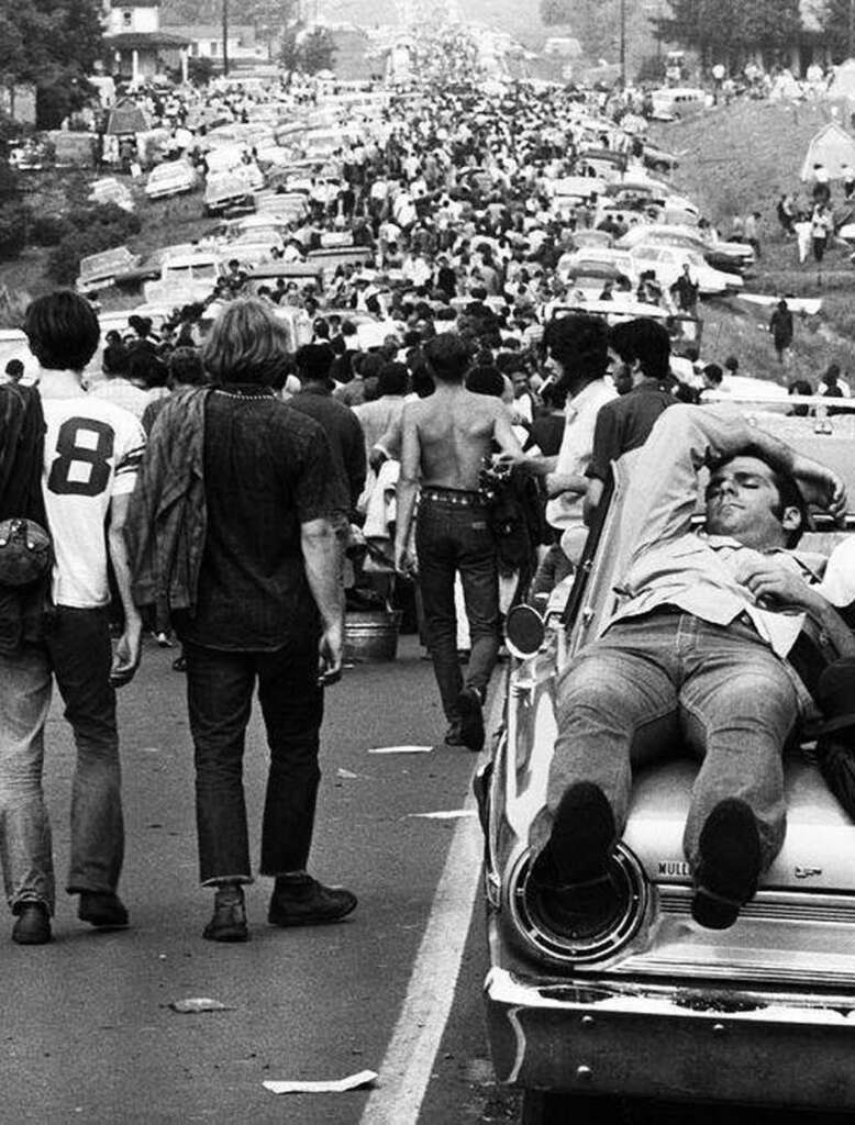 Black and white photo of a large crowd walking on a road filled with cars. A man is lying on the hood of a car in the foreground, wearing a jacket and pants. People are casually dressed, with some shirtless, moving away from the camera.