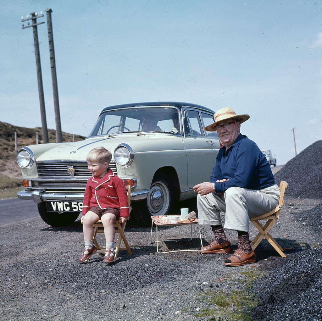 An elderly man in a straw hat and a young boy sit on folding chairs by a roadside. A light green vintage car is parked behind them. They appear to be enjoying a break, with a simple meal on a small table, under a clear blue sky.