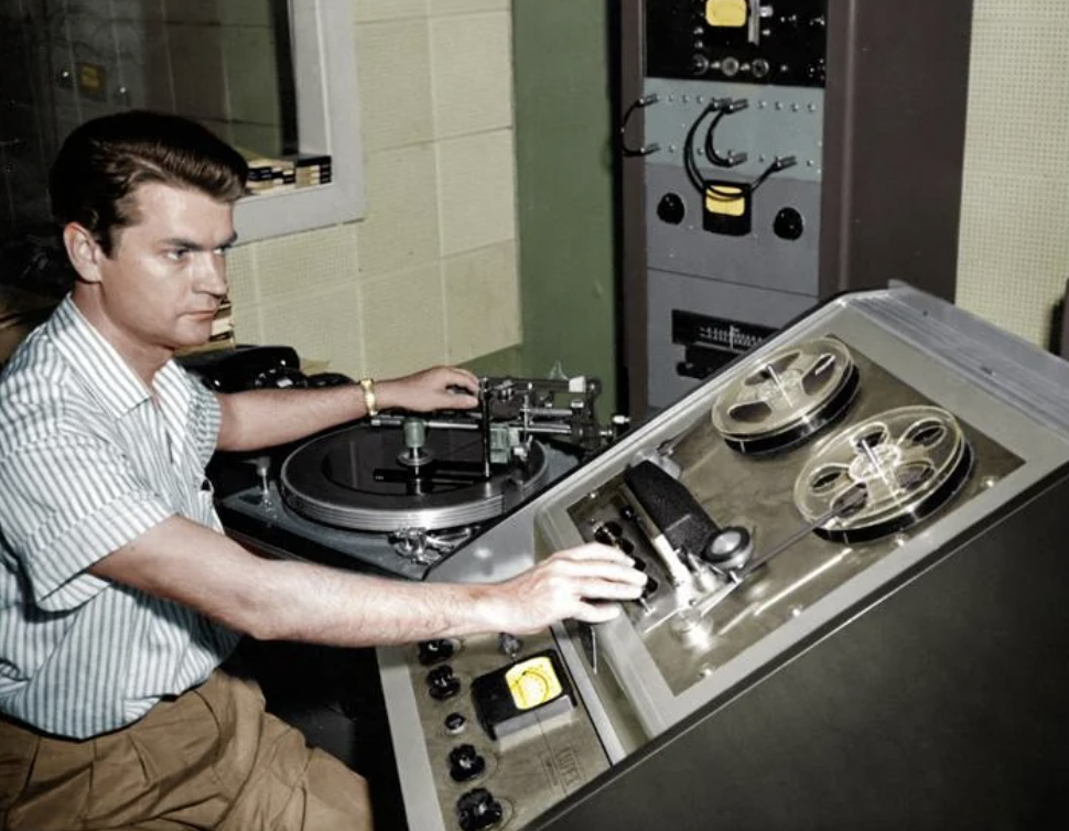 A person operates vintage recording equipment, adjusting dials and handling reel-to-reel tape in a studio. They wear a striped shirt and have a focused expression. The equipment includes reels, a turntable, and various controls.