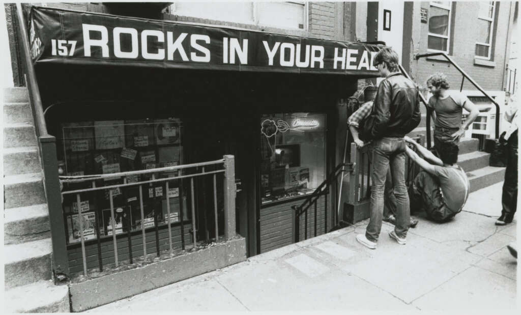 Black and white photo of people outside "Rocks in Your Head," a record store with a sign displaying the name. The entrance is below street level with visible steps. A group of people are sitting and standing near the entrance.