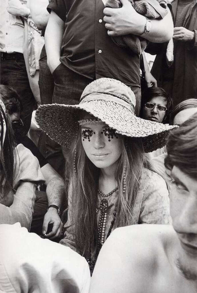 A woman with long hair sits among a crowd. She is wearing a wide-brimmed straw hat and has large decorative eyelashes. The black-and-white image captures the fashion and atmosphere of the scene.