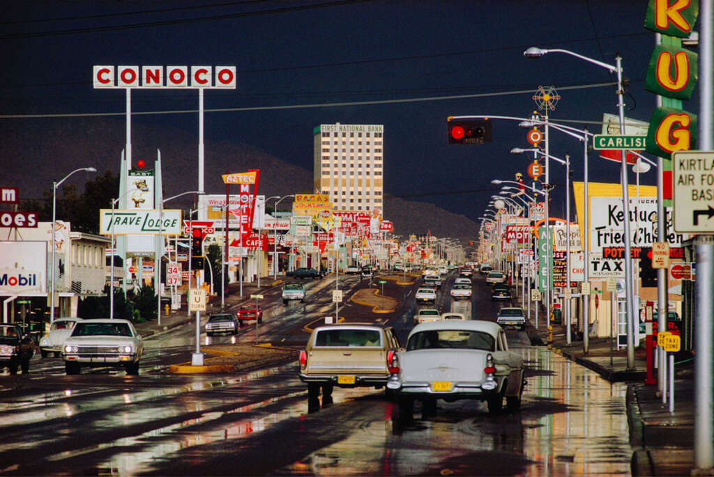 A vibrant street scene depicting a busy road lined with colorful vintage neon signs and billboards for various businesses. Classic cars drive on the wet street under a cloudy sky, with mountains visible in the background.