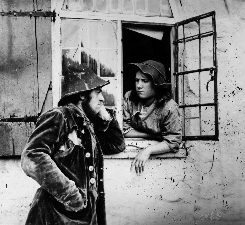 Two individuals in rustic attire converse outside a window. One leans against the windowsill, while the other stands with a hand on their face, both wearing hats and old-fashioned clothing. The window is partially open, revealing a simple interior.