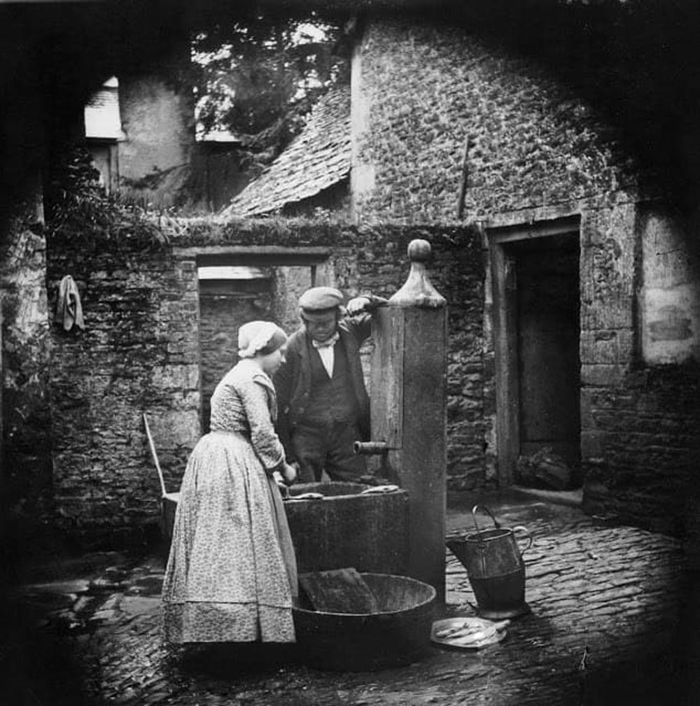 A vintage photo shows a man and woman in old-fashioned clothing gathering water from a stone fountain in a cobblestone courtyard. A bucket sits on the ground nearby, and stone buildings surround the scene.