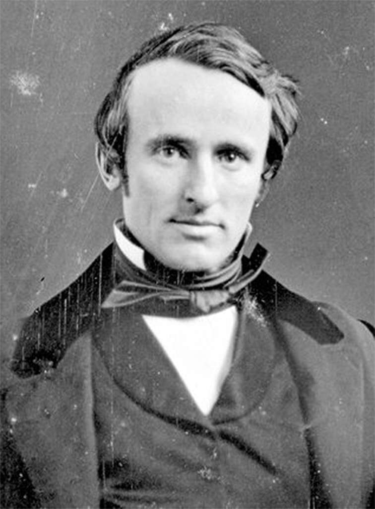 A vintage black and white photograph of a man in 19th-century attire. He wears a dark coat, white shirt, and a bow tie, with neatly combed hair. The image has a slightly faded and scratched appearance, typical of older photographs.