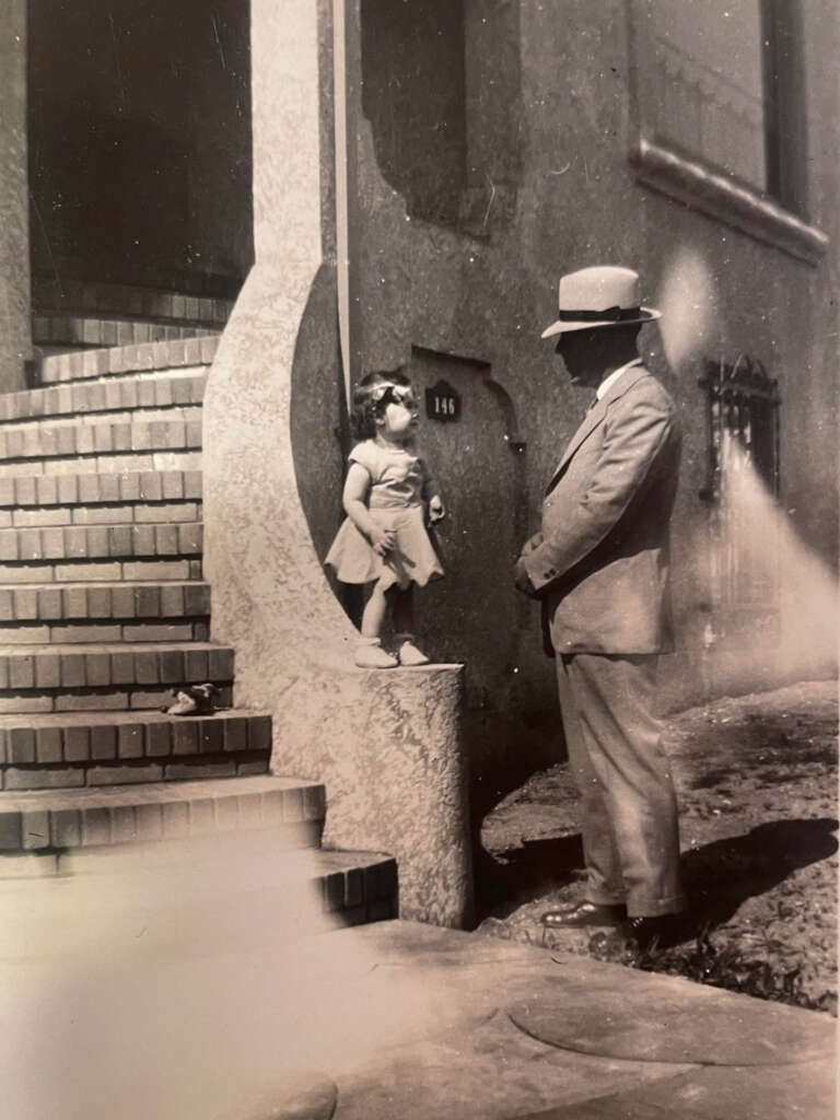 A vintage photo of a man in a suit and hat standing on a sidewalk, looking up at a young girl in a dress who is standing on the brick steps of a building. The building has an arched entrance and a "146" sign.