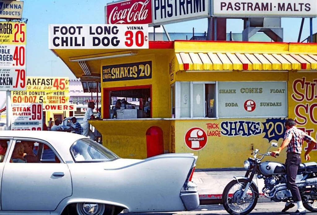 A vintage scene with a yellow and red food stand offering hot dogs, shakes, and pastrami. A classic white car and a motorcycle are parked in front. The stand is adorned with large, bold signage advertising food items and prices.