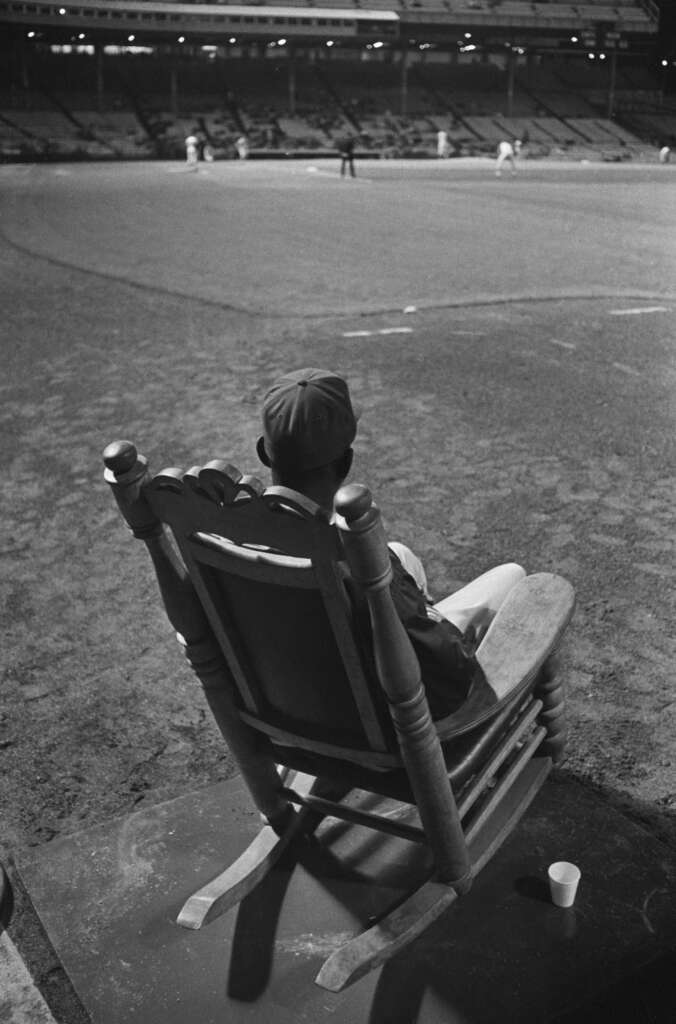 A person in a baseball uniform sits in a rocking chair at the edge of a baseball field, facing the action. The stadium is illuminated, with players and spectators visible in the background. A paper cup is on the ground nearby.