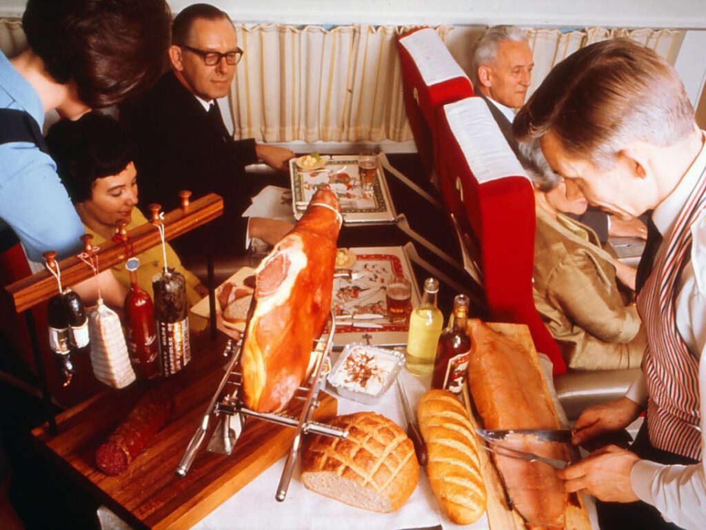 Passengers on an airplane enjoy a gourmet meal service. A flight attendant serves food while a man slices cured meats and bread. The table is set with various condiments. The cabin features red seats and a curtain.