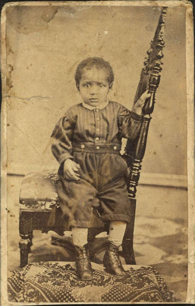 A sepia-toned vintage photograph of a child sitting on a chair. The child is wearing a dark, long-sleeved outfit with buttons and high-top boots. The setting includes a patterned rug and a plain backdrop.