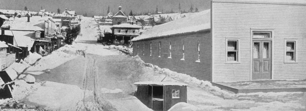 Black and white photo of a snow-covered street in a small town. Wooden buildings line the street, one with "Livery Stable" signage visible. Tracks lead up the hill. The scene has a historical and quiet winter atmosphere.