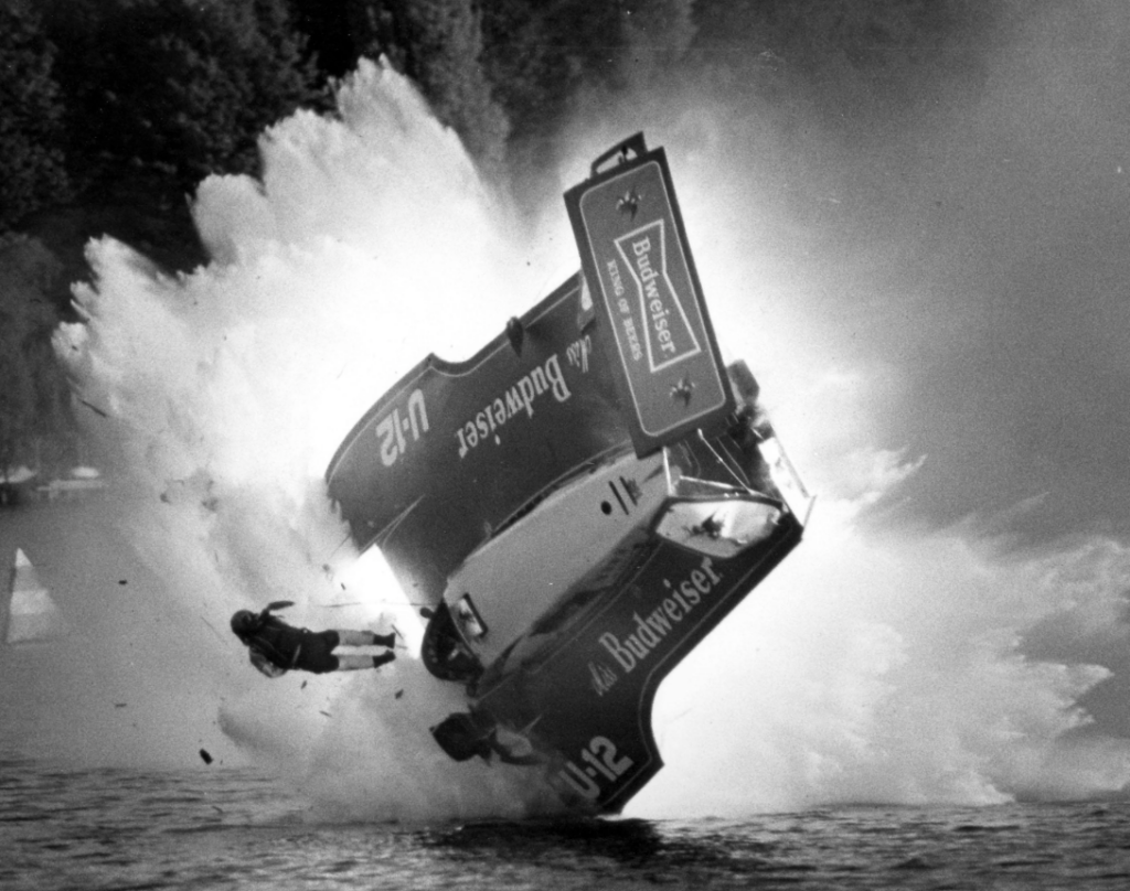 A speedboat labeled "Budweiser" flips dramatically during a race, with a large splash of water around it. A person appears to be ejected into the air. Trees and water are in the background. The photo is in black and white.