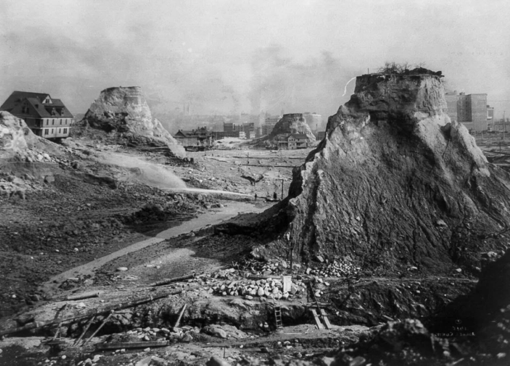 Black and white photo of an eroded landscape in Seattle from the early 20th century. Large dirt mounds and excavated surfaces are visible, with a few scattered buildings in the background and a hazy skyline.