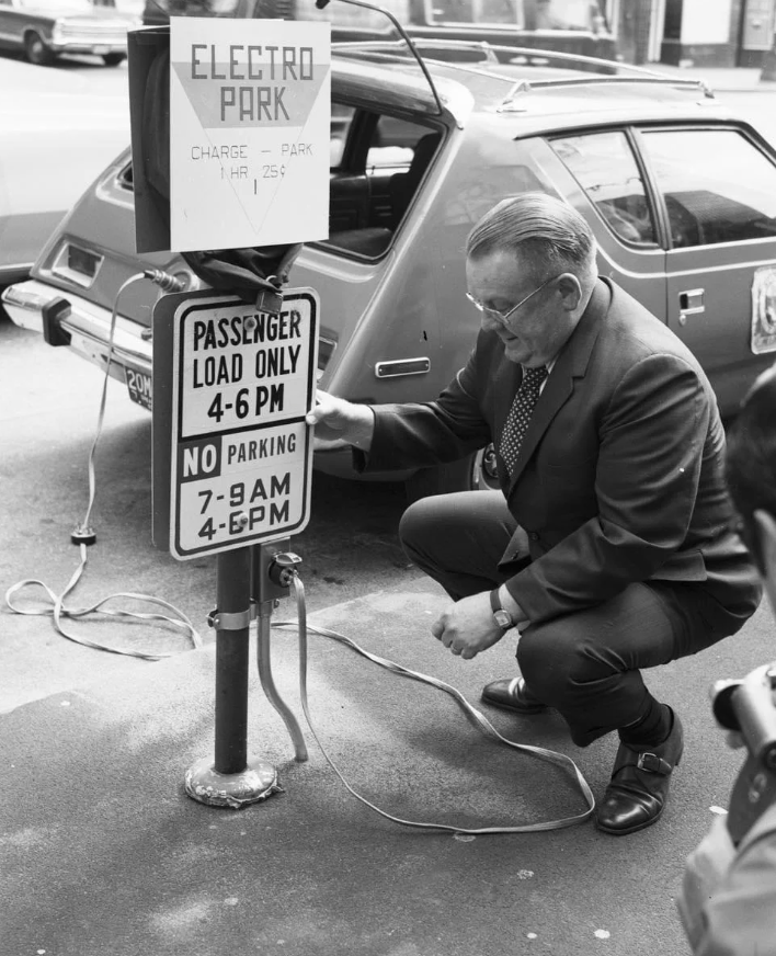 A man in a suit kneels beside an early electric car charging station labeled "Electro Park" with a sign showing parking and charging hours. A vintage car is parked nearby, and cables connect the car to the station.