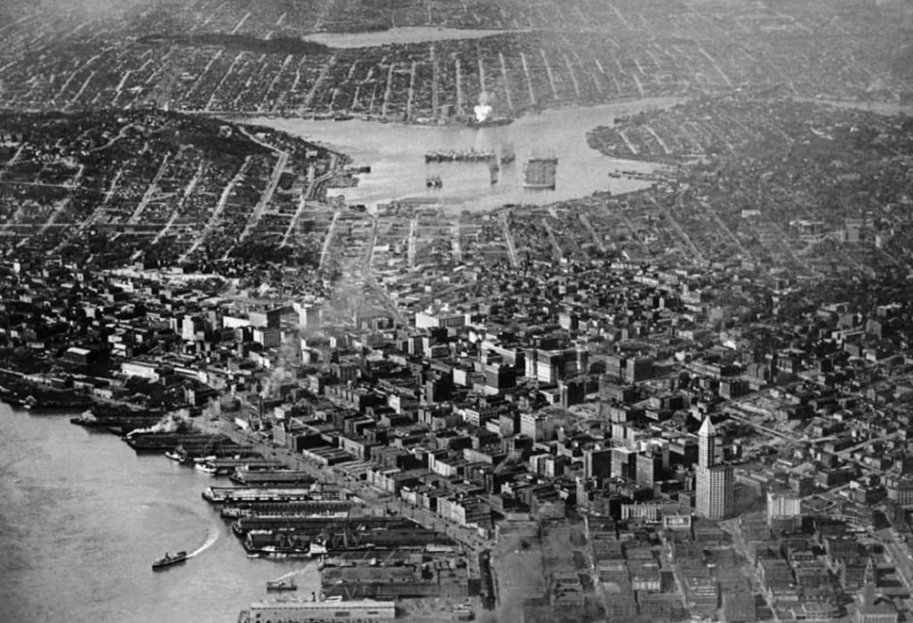 Aerial black and white photo of a city with dense urban buildings by the waterfront. A large harbor with ships is visible, and a network of streets stretches into the distance, bordered by a river or inlet.