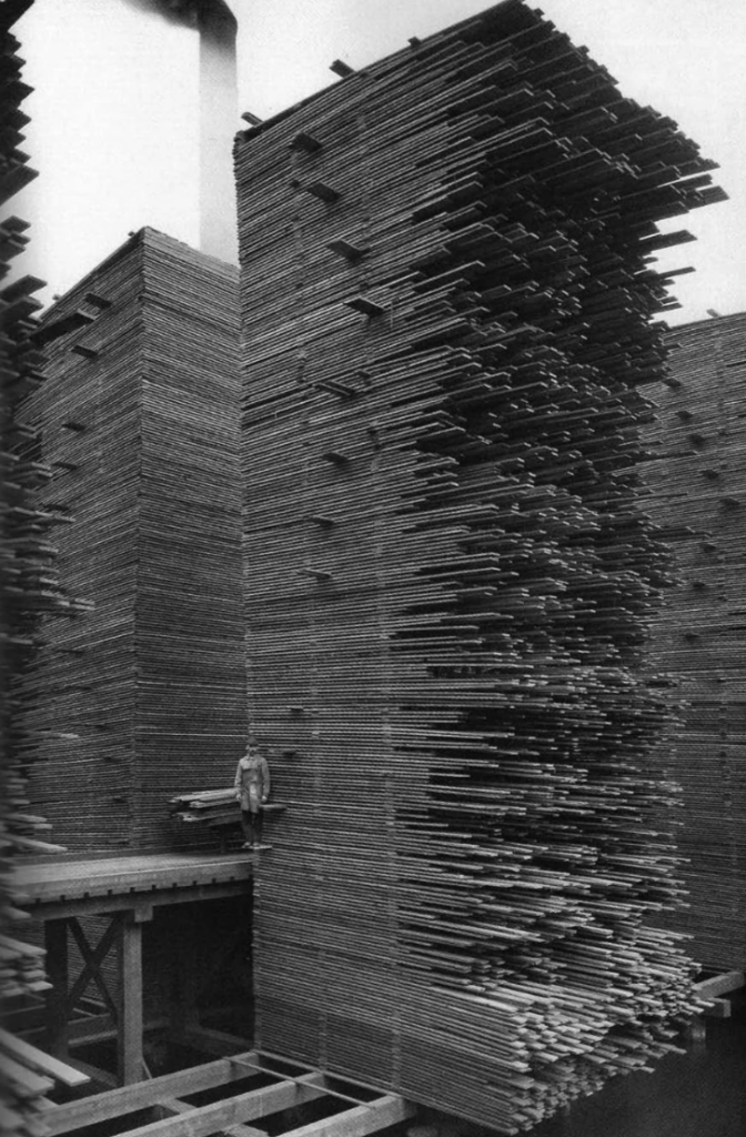 A black-and-white photo showing a man standing next to a towering stack of wooden planks. The planks are piled vertically to an impressive height, emphasizing the scale compared to the small figure standing beside them.