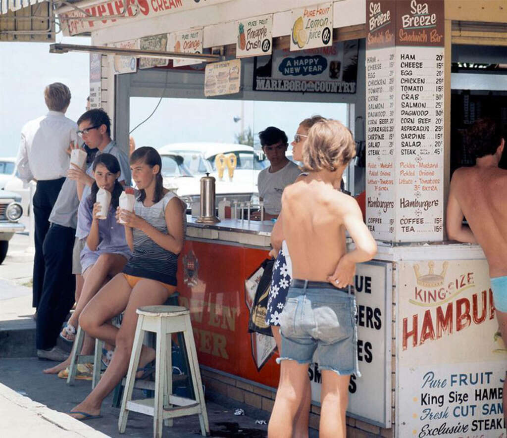 People gather outside a vintage seaside snack bar. Four young adults sit on stools, drinking milkshakes, while a boy in shorts and no shirt stands nearby. A man in sunglasses leans against a wall, and cars are parked in the background.