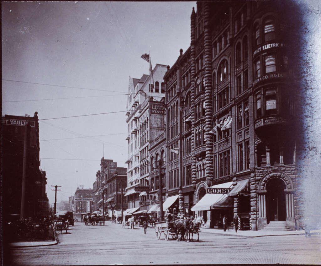 A vintage photo of a bustling city street scene from the late 19th or early 20th century. Horse-drawn carriages move along a cobblestone road lined with ornate brick buildings and storefronts. People are walking on the sidewalks.