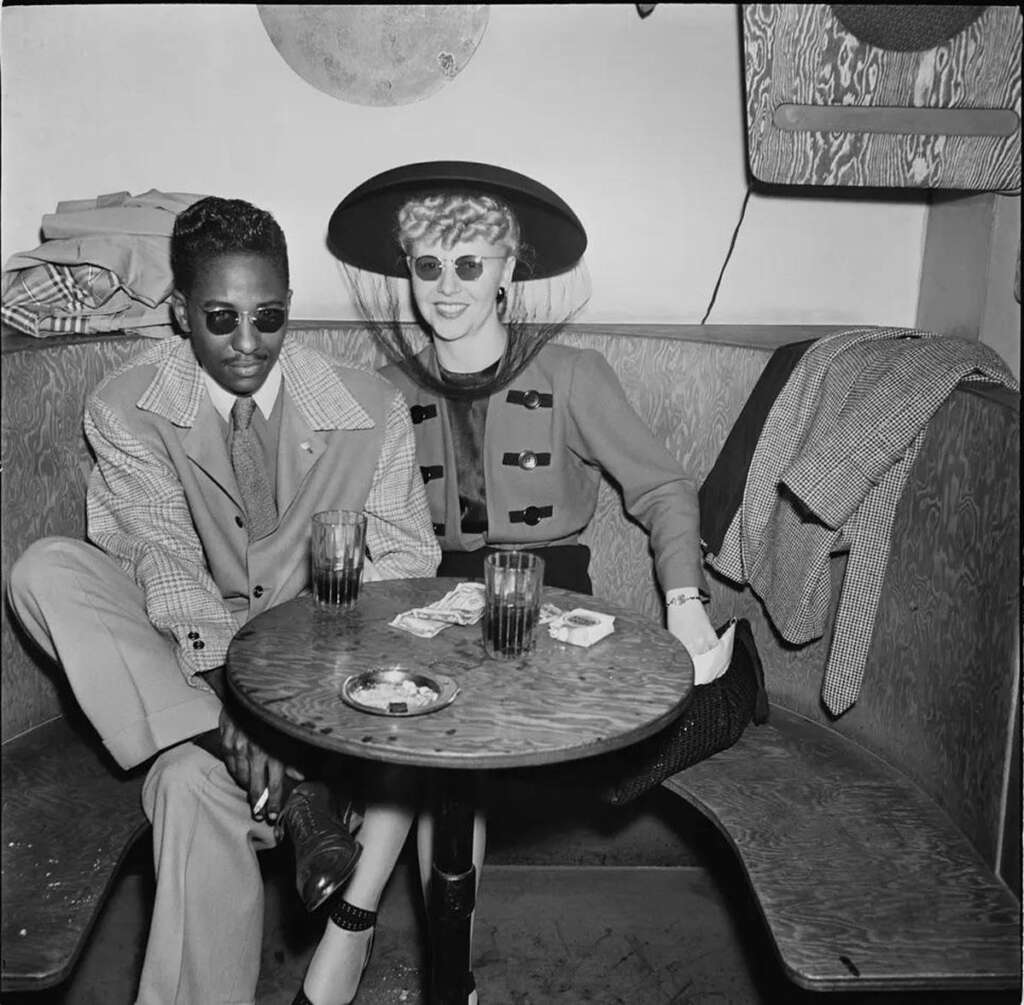 A man and woman sit together in a booth at a café. The man wears a suit and sunglasses, while the woman is in a stylish dress and wide-brimmed hat. Drinks and cigarettes are on the table. Retro, black-and-white photo.