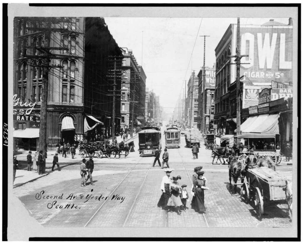 Historic black-and-white photo of Second Ave and Yesler Way in Seattle, featuring streetcars, pedestrians, and horse-drawn carriages amidst tall buildings and signs for businesses, including a prominently displayed Owl Cigar ad.