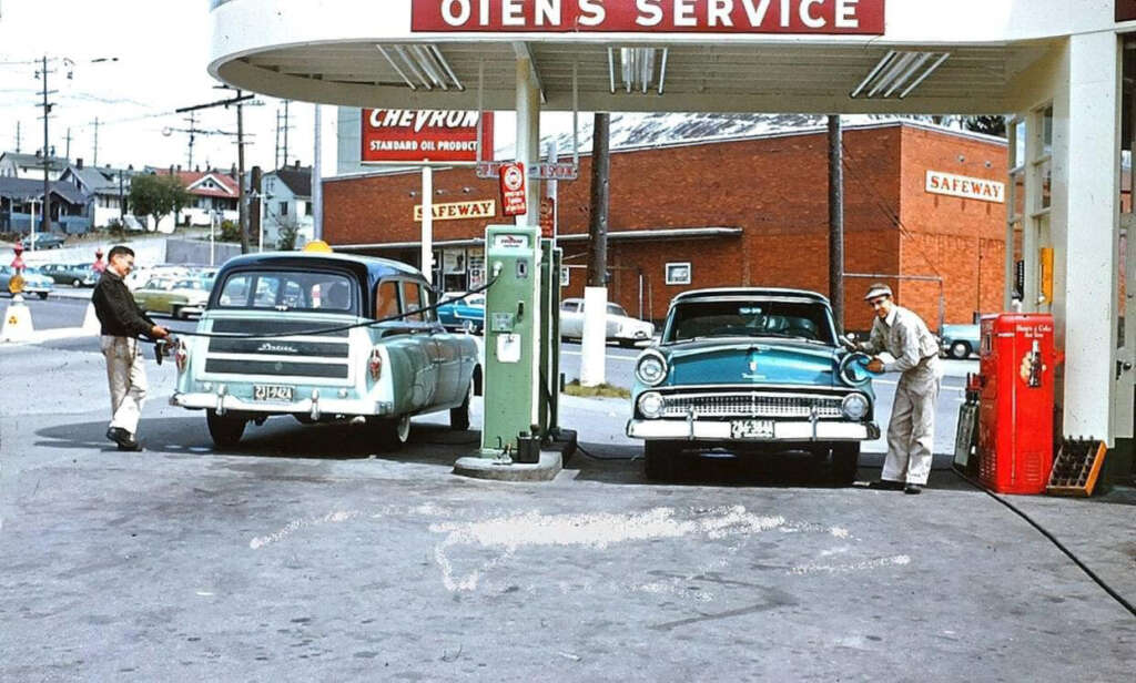 A vintage gas station scene with two classic cars being refueled. An attendant in a white uniform fills the tank of a turquoise car. Another person stands by the open door of a light blue car. Buildings and signs are in the background.