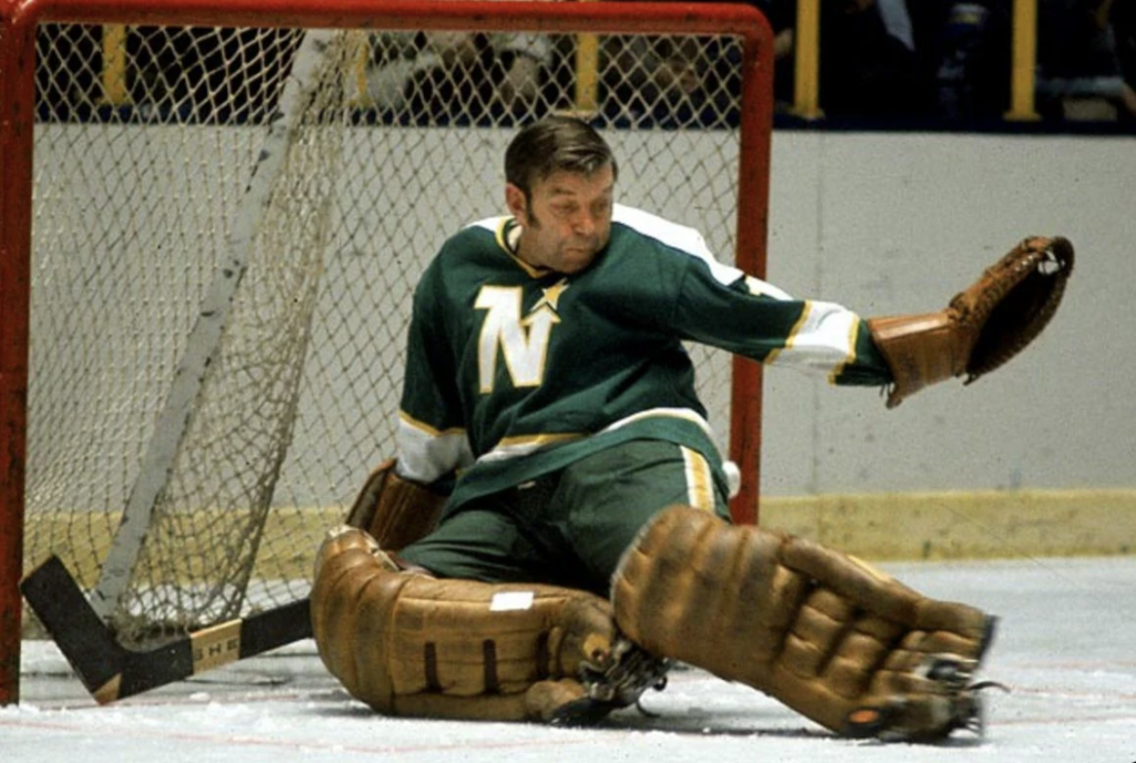 A hockey goalie in green gear is making a save in front of the goal. He wears vintage brown pads and gloves, with the letter 'N' prominent on his jersey. The net and hockey rink boards are visible in the background.