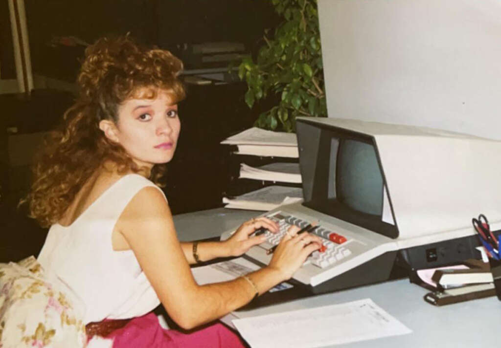 A woman with curly hair sits at a vintage computer, typing. She wears a white top and pink skirt, surrounded by papers and a desk with office supplies. A plant is visible in the background.