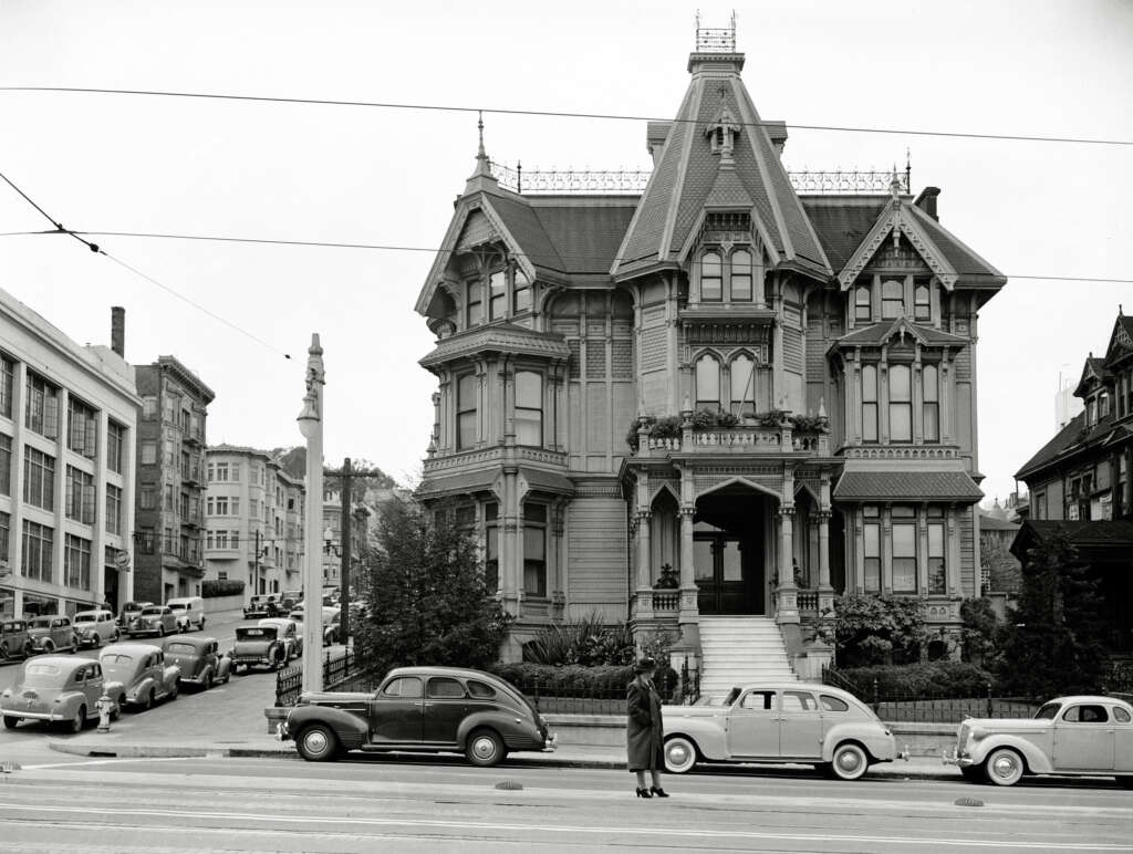 A black-and-white photo of a Victorian-style house with intricate detailing, located on a street corner. Several vintage cars are parked along the street, and a person stands in front, facing the house. Trees surround the area, with buildings nearby.