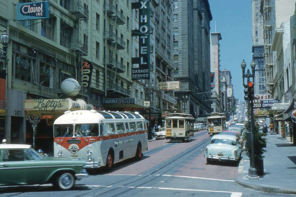 A bustling 1950s urban street scene with vintage cars, a trolleybus, and cable cars. Tall buildings line the street, featuring signs for a hotel, shops, and a garage. Pedestrians walk along the sidewalks, and traffic lights control the intersection.