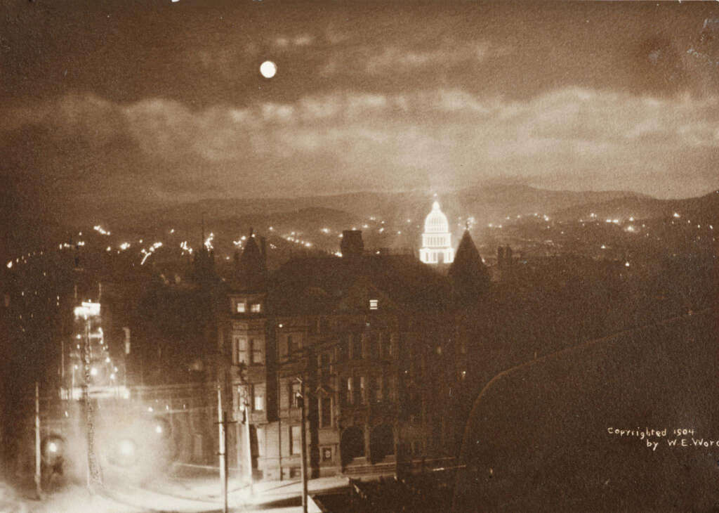 A sepia-toned nighttime cityscape from 1900 shows a brightly lit, domed building in the distance. Streets are illuminated by streetlights, with a prominent moon in the cloudy sky above. Multiple buildings and hills are visible in the background.