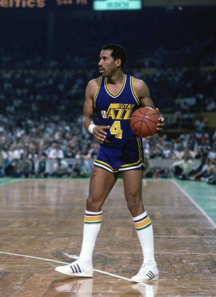 A basketball player in a blue and yellow Utah Jazz uniform is dribbling a basketball on a wooden court. He's wearing white socks and sneakers. The audience is visible in the blurred background.