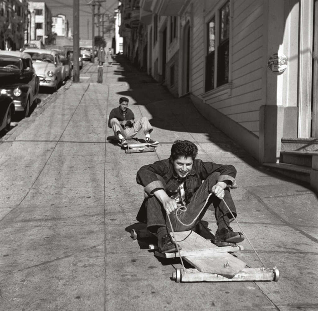 Two people are riding makeshift wooden carts downhill on a steep city street. The road is lined with parked cars and buildings. The front rider appears excited, gripping a rope, while the second person follows behind.