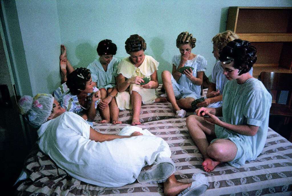 Seven women with hair rollers sit and relax on a bed in a bright room. They are engaged in conversation and activities such as reading and doing nails. The room has a minimalist design with a wooden chair and an empty shelving unit.