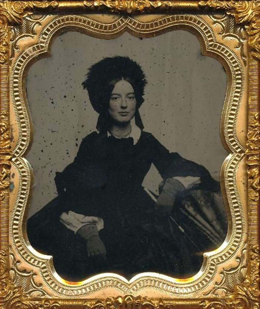 A vintage photograph of a woman in a dark dress and a large feathered hat, sitting with one arm on a surface. The image is enclosed in an ornate, gold frame. The woman gazes directly at the camera with a composed expression.