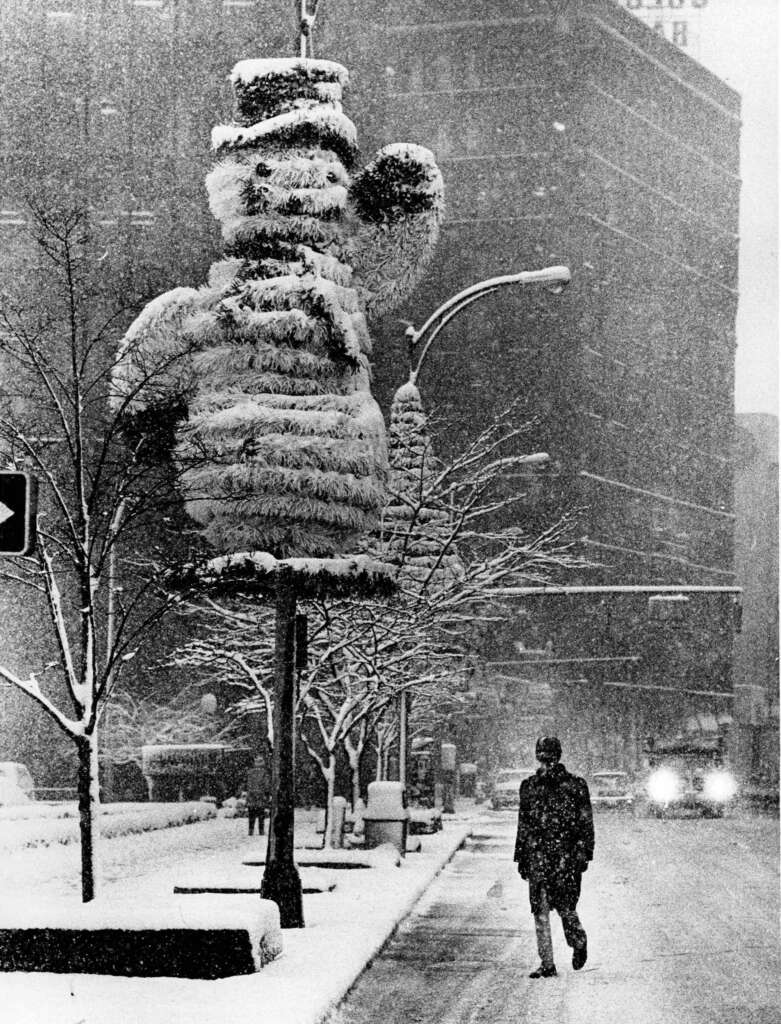 A black-and-white image of a snowy city street. A person walks along the sidewalk. Large snow-covered figures shaped like snowmen hang from streetlights. A vehicle with headlights on approaches in the background. Snow blankets the scene.