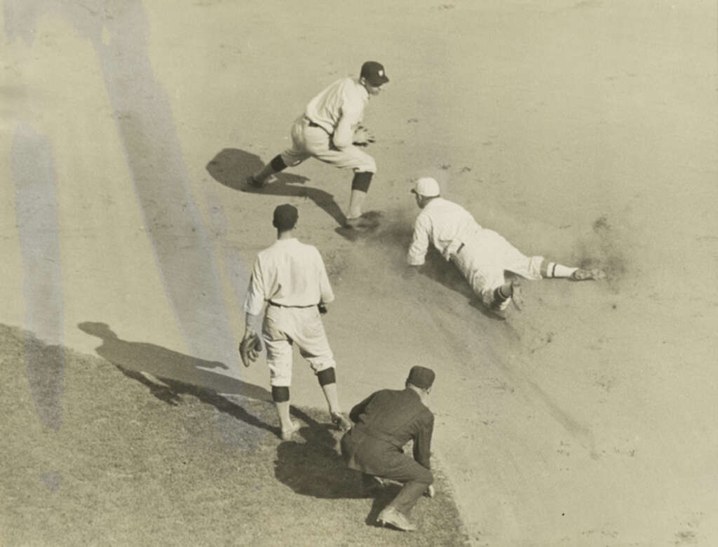 A sepia-toned image shows a baseball game scene from above. A player slides into a base as the baseman reaches for him. Two other players and an umpire stand nearby. Dust surrounds the area, suggesting the action of the slide.