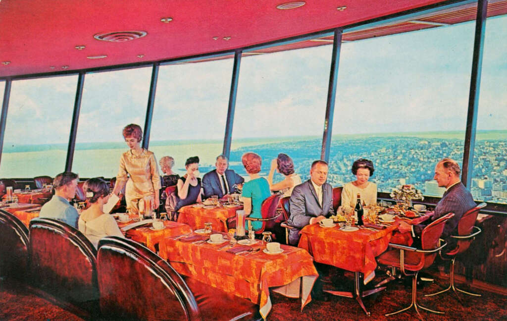 A group of people dine at an elevated restaurant with large windows overlooking a cityscape and body of water. The interior has red decor, and a server attends to them. The tables are covered with red tablecloths.