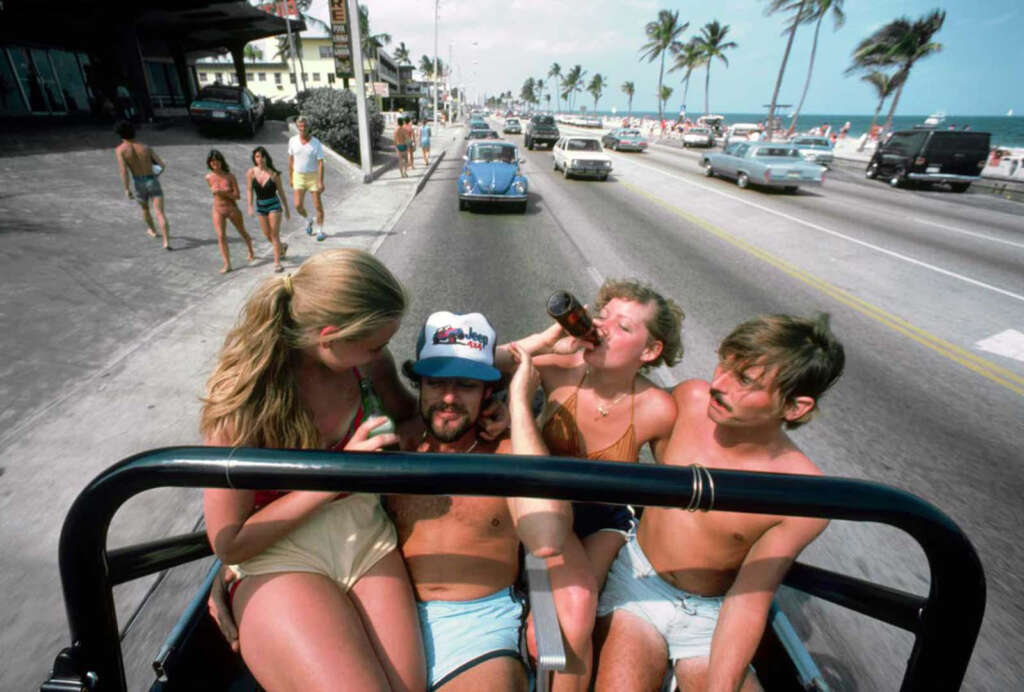 Four people in swimsuits ride in a convertible on a sunny beach road. Two women drink from bottles, and a man in a cap is seated in the middle. Palm trees line the street, and beachgoers and cars are visible in the background.