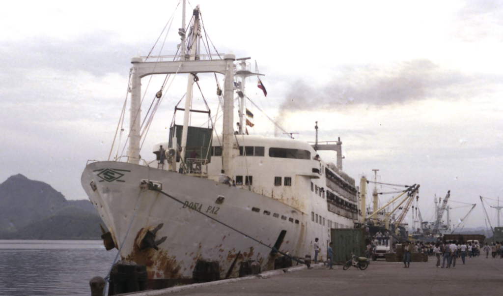 A large, rusted white passenger ship named "Babael Paz" is docked at a port. Smoke is emitting from its funnel. People are gathered near the dock, and cranes are visible in the background under a cloudy sky.