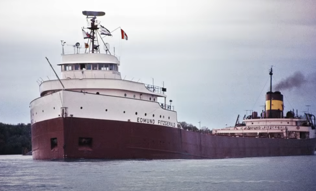 A large cargo ship named Edmund Fitzgerald sails on a calm body of water. The vessel has a white upper structure and a red hull. Smoke is visible from the smokestack, and a flag is raised on the mast. The sky is overcast.