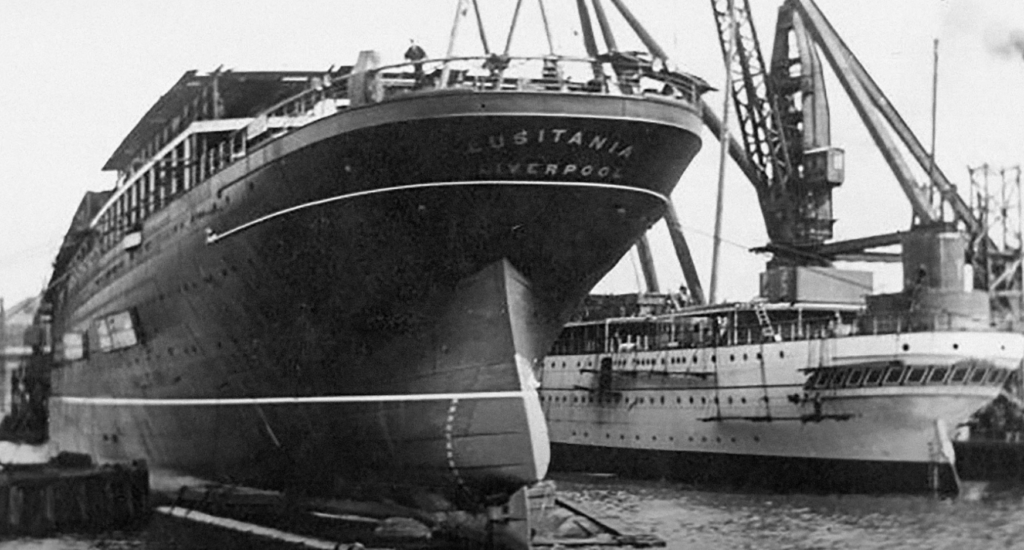 Black and white photo of the RMS Lusitania, with its name visible on the stern, docked next to another large ship. Cranes are present in the background, indicating an industrial shipyard setting.