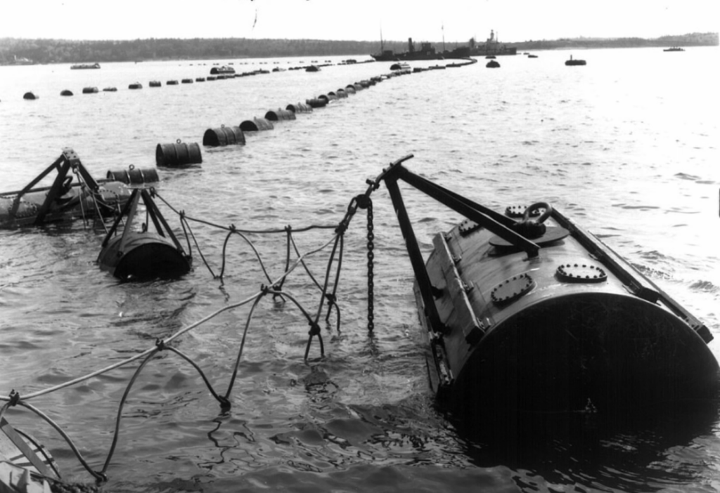 A black and white image showing a line of large, cylindrical marine navigation buoys connected by cables, floating in water. The background reveals a distant shoreline and boats on the horizon.