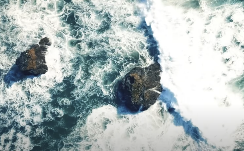 Aerial view of waves crashing against two large rocks in the ocean. The water is a mix of deep blue and foamy white, creating swirling patterns around the dark, jagged rocks.