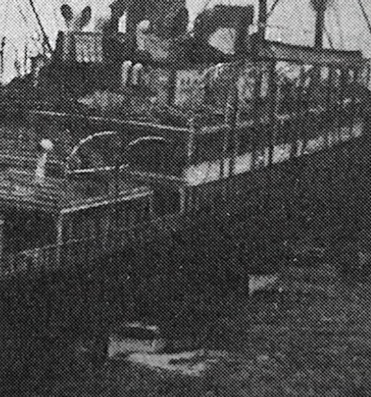 A vintage black and white photo of a wooden ship on water, likely from the steam era. The vessel has multiple decks with visible rails and chimneys emitting smoke, suggesting it's in operation. The image has a grainy texture.