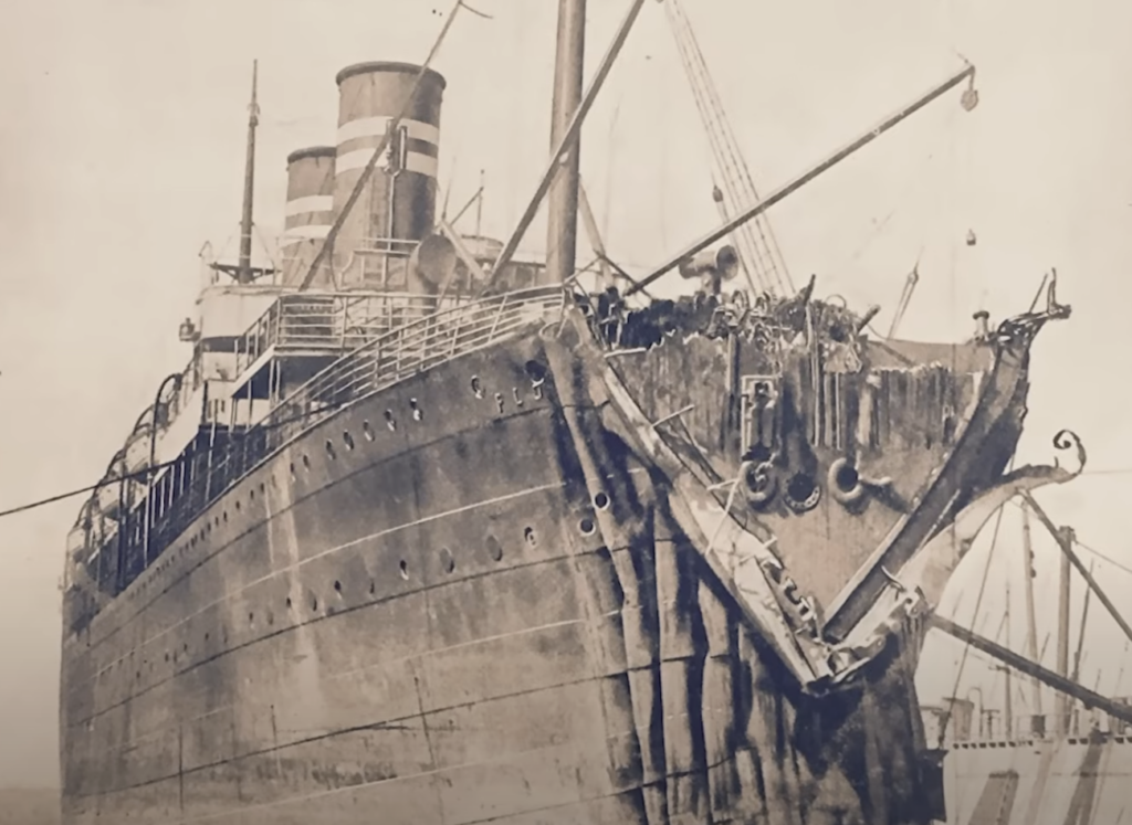 A vintage photograph of a large ship displaying a severely damaged bow. The vessel shows visible dents and structural damage at the front. The ship has a tall, striped smokestack and is docked at a port. Cranes and ropes are visible in the background.