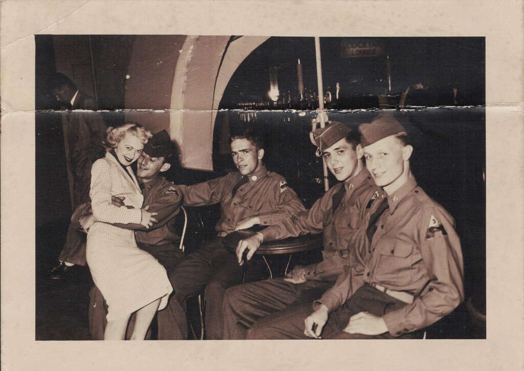 A group of four soldiers in uniform are seated at a table. One soldier has a woman sitting on his lap, smiling. They are in a room with arched windows and city lights visible outside. The atmosphere is relaxed and cheerful.