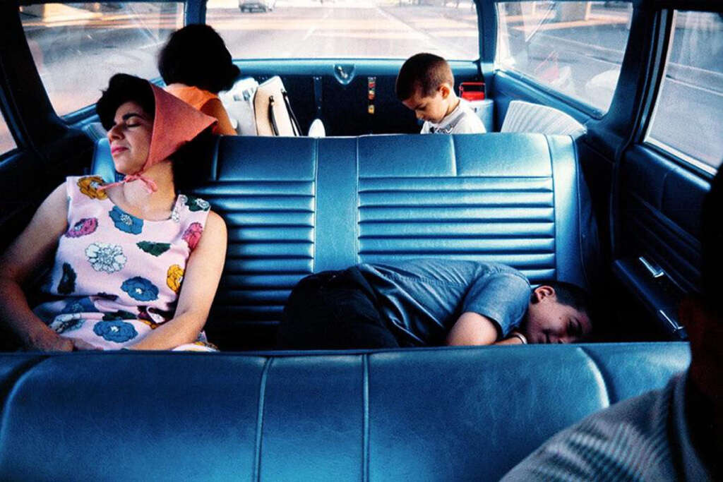 A woman with a pink hat rests against the car door in the front seat, while two children in the back seat are asleep. A third child in the back looks down, and a driver is partially visible. The car interior is blue.