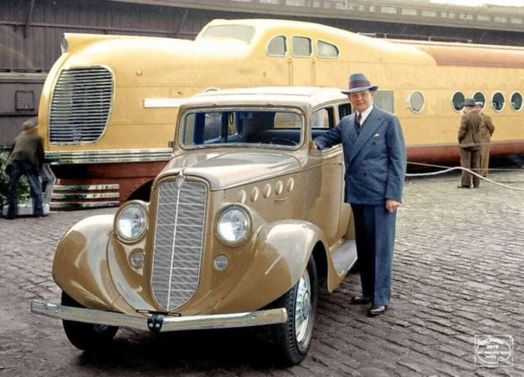 A man in a blue suit and hat stands beside a vintage brown car. Behind them, there's a large, streamlined, yellow train carriage. Other people are visible in the background. The setting appears to be an outdoor display area.