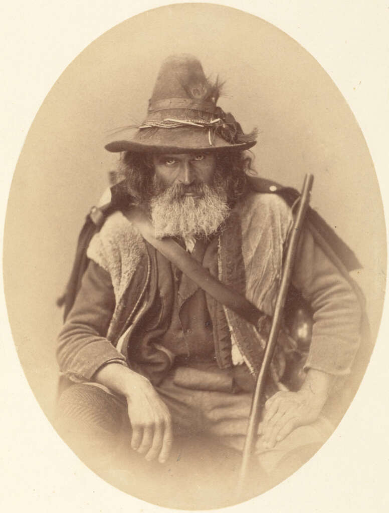 A sepia-toned vintage photograph of an older man with a long, bushy white beard and hair. He wears a tattered wide-brimmed hat adorned with feathers and holds a long stick, dressed in layered, rough garments. The photo has an oval border.