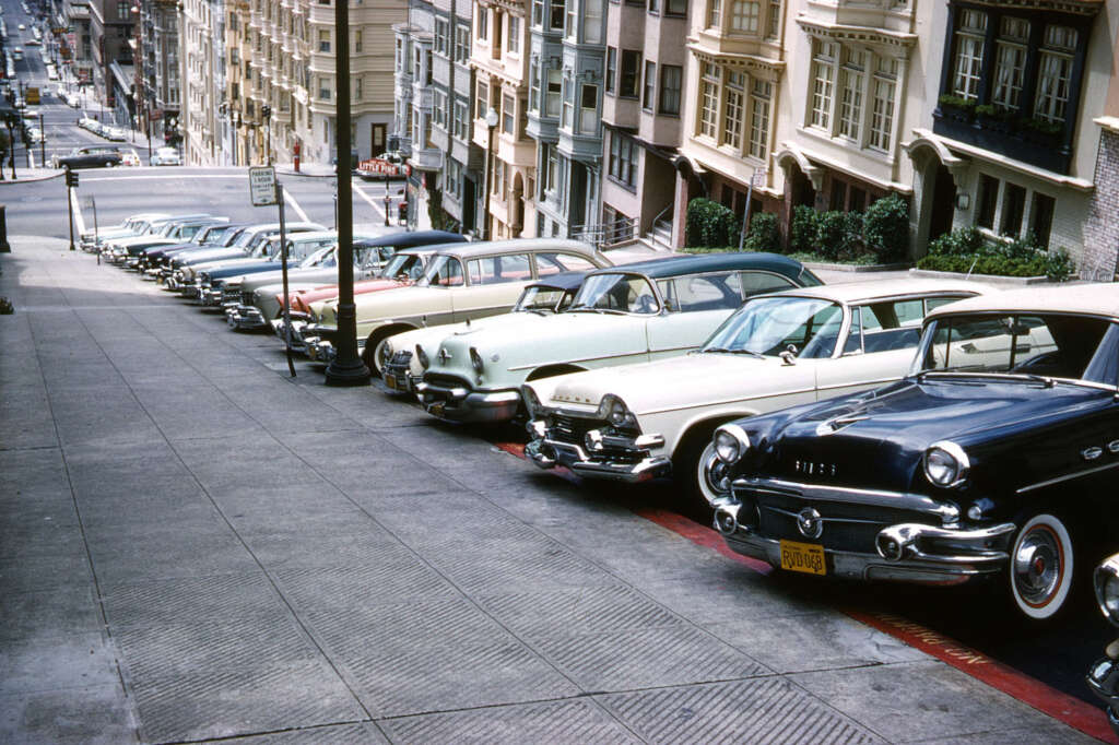 A row of 1950s cars parked diagonally along a steep, urban street lined with multi-story buildings. The street slopes downwards, creating a dramatic perspective. The buildings are detailed and closely packed.